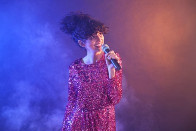 Beautiful young woman with microphone singing on color background in neon lights and smoke