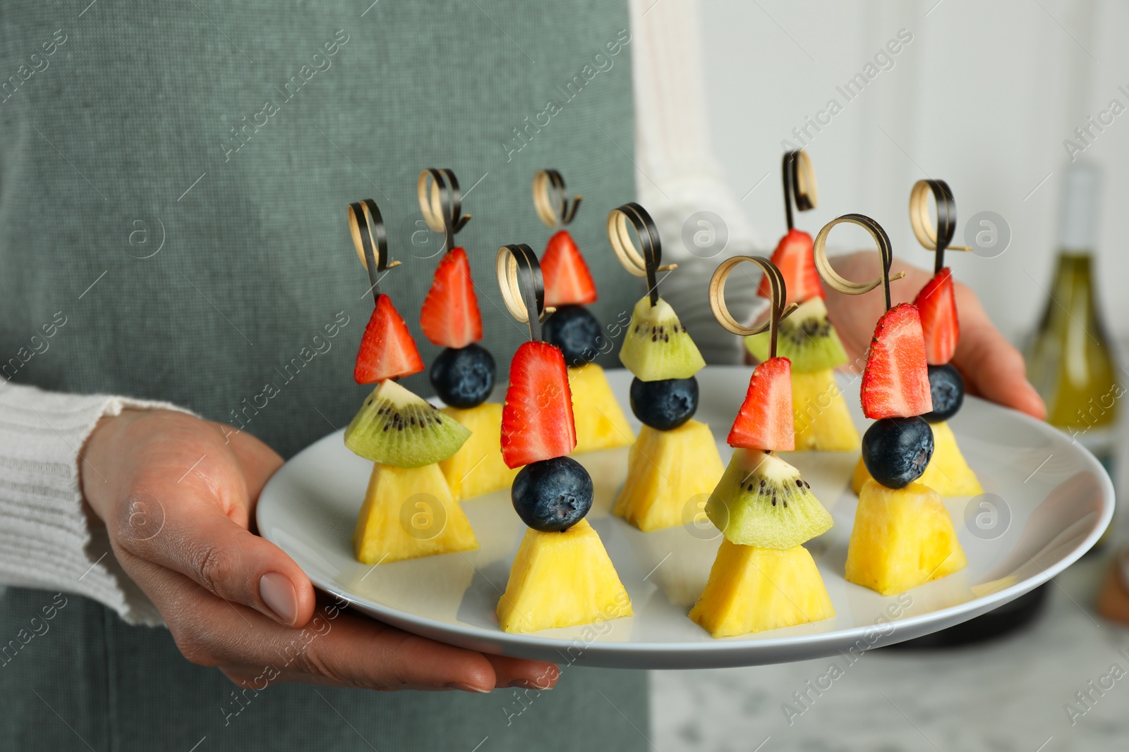 Photo of Woman holding plate of tasty canapes with pineapple, kiwi and berries indoors, closeup