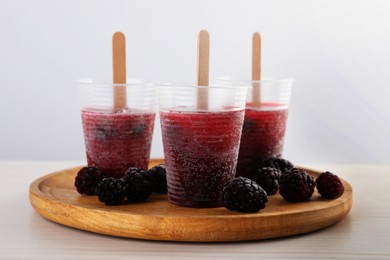 Photo of Tasty blackberry ice pops in plastic cups on white wooden table. Fruit popsicle