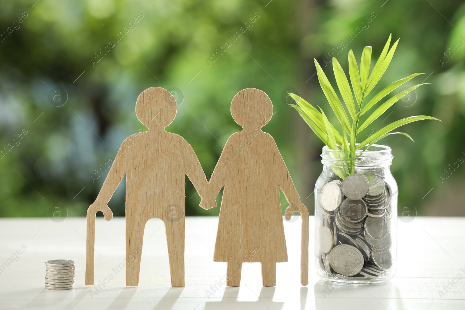 Photo of Pension savings. Figure of senior couple, jar with coins and twig on white table against blurred green background