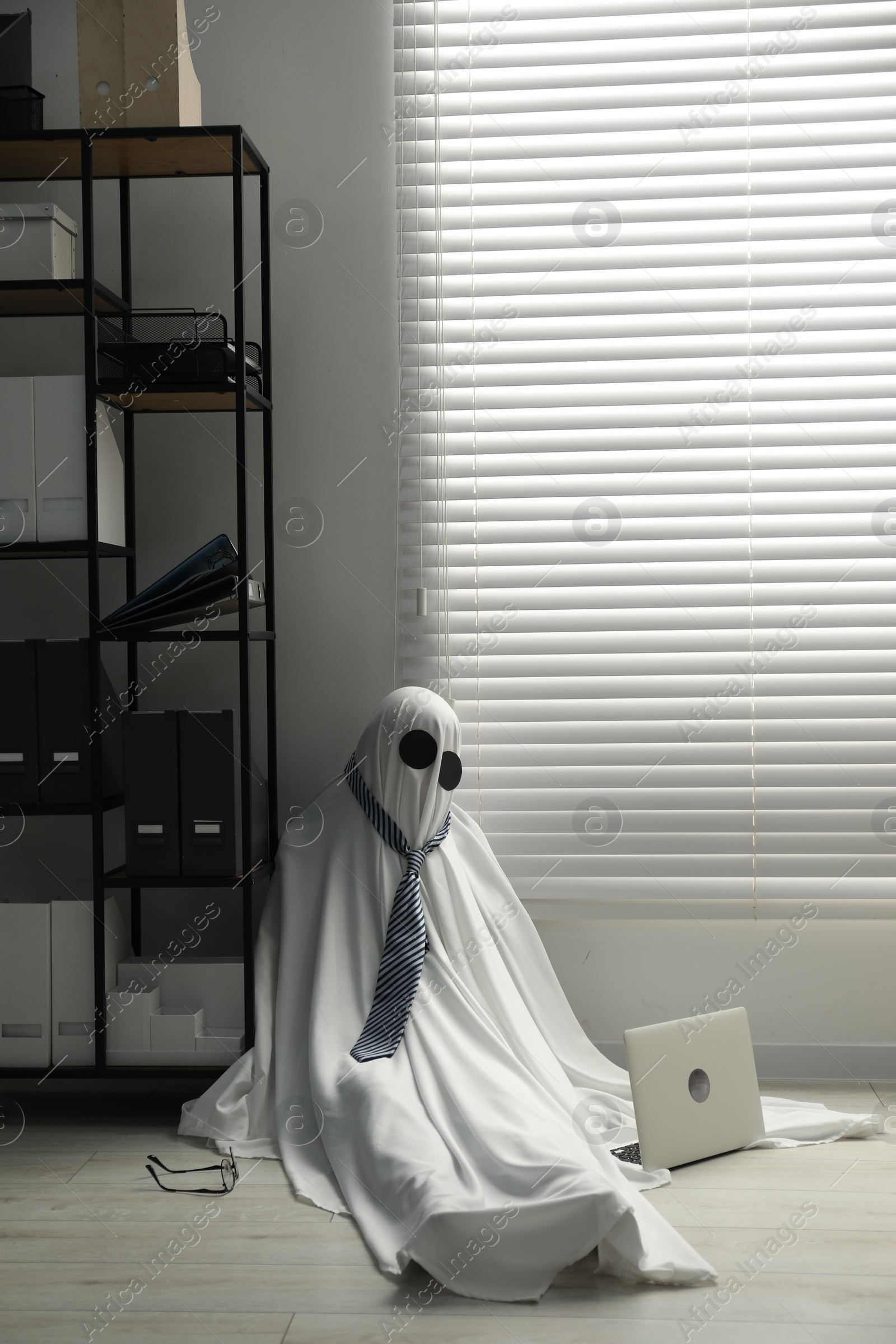 Photo of Overworked ghost. Man in white sheet with laptop on floor in office