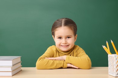 Photo of Happy little school child sitting at desk with books near chalkboard