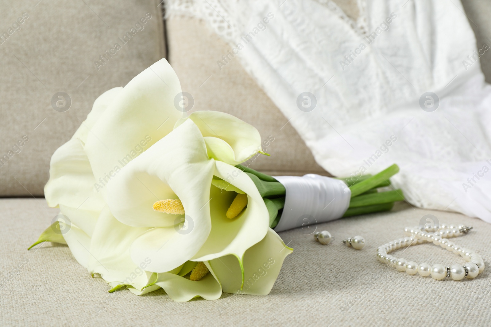 Photo of Beautiful calla lily flowers tied with ribbon, wedding dress and jewelry on sofa