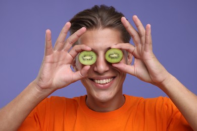 Photo of Smiling man covering eyes with halves of kiwi on violet background, closeup