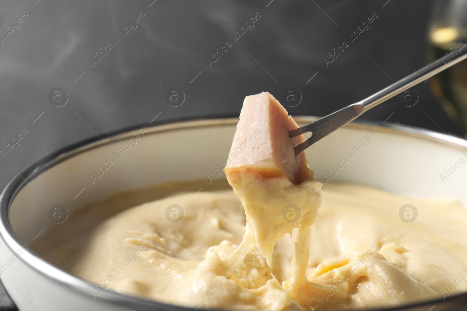 Photo of Dipping piece of ham into fondue pot with melted cheese on grey background, closeup