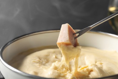 Photo of Dipping piece of ham into fondue pot with melted cheese on grey background, closeup