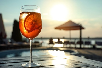 Glass of fresh summer cocktail on wooden table near swimming pool outdoors at sunset. Space for text