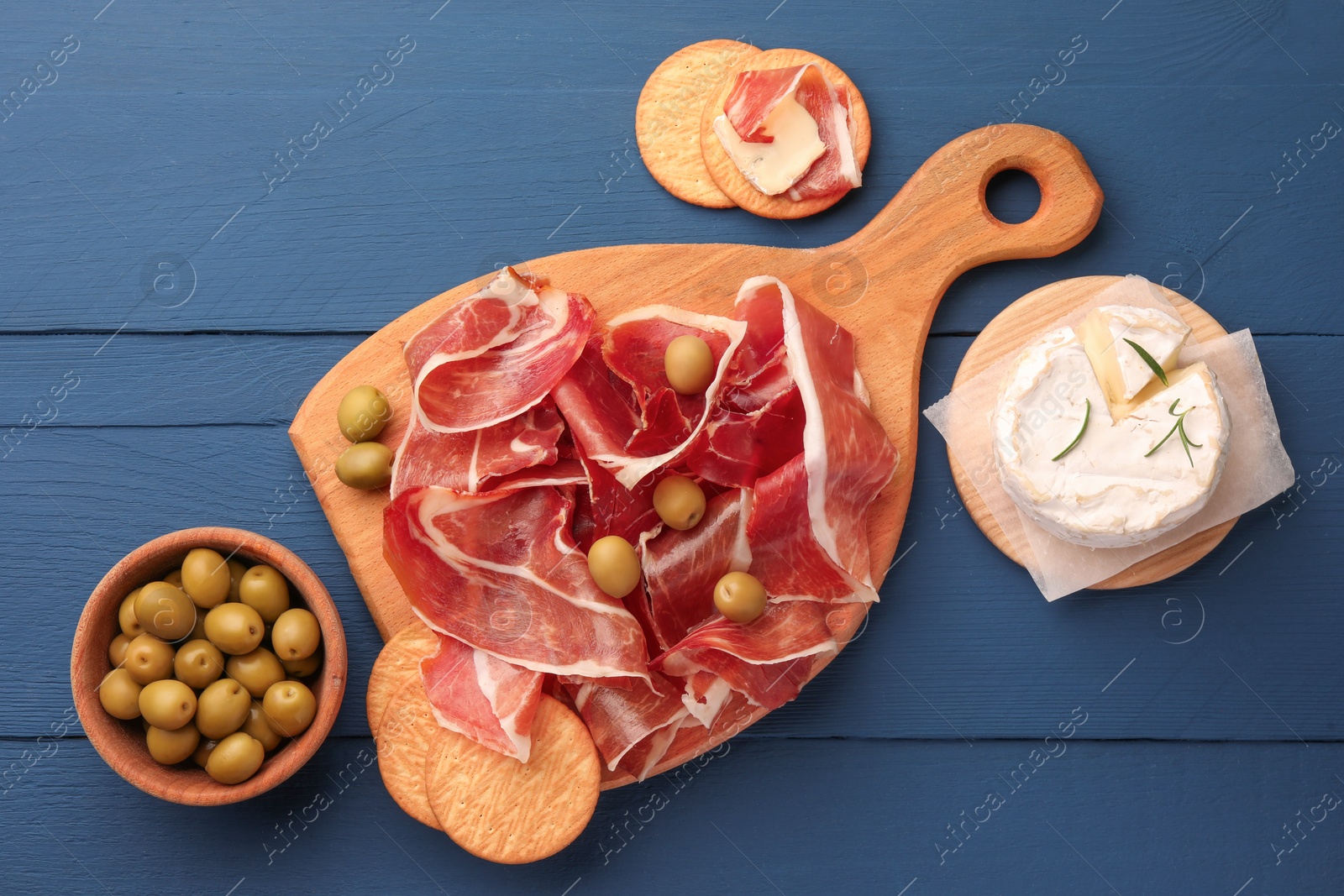 Photo of Slices of tasty cured ham, olives and cheese on blue wooden table, flat lay