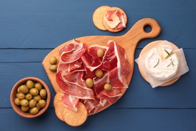 Slices of tasty cured ham, olives and cheese on blue wooden table, flat lay