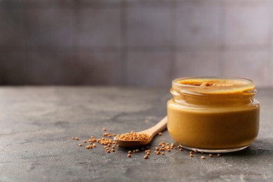 Photo of Tasty mustard sauce in jar and spoon with dry seeds on grey textured table, space for text