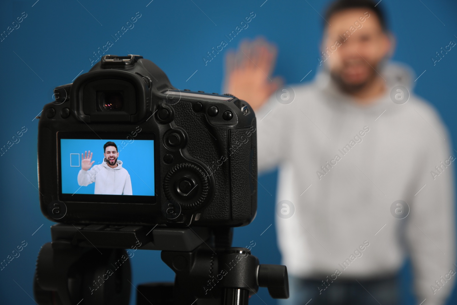 Photo of Young blogger shooting video with camera against blue background, focus on screen