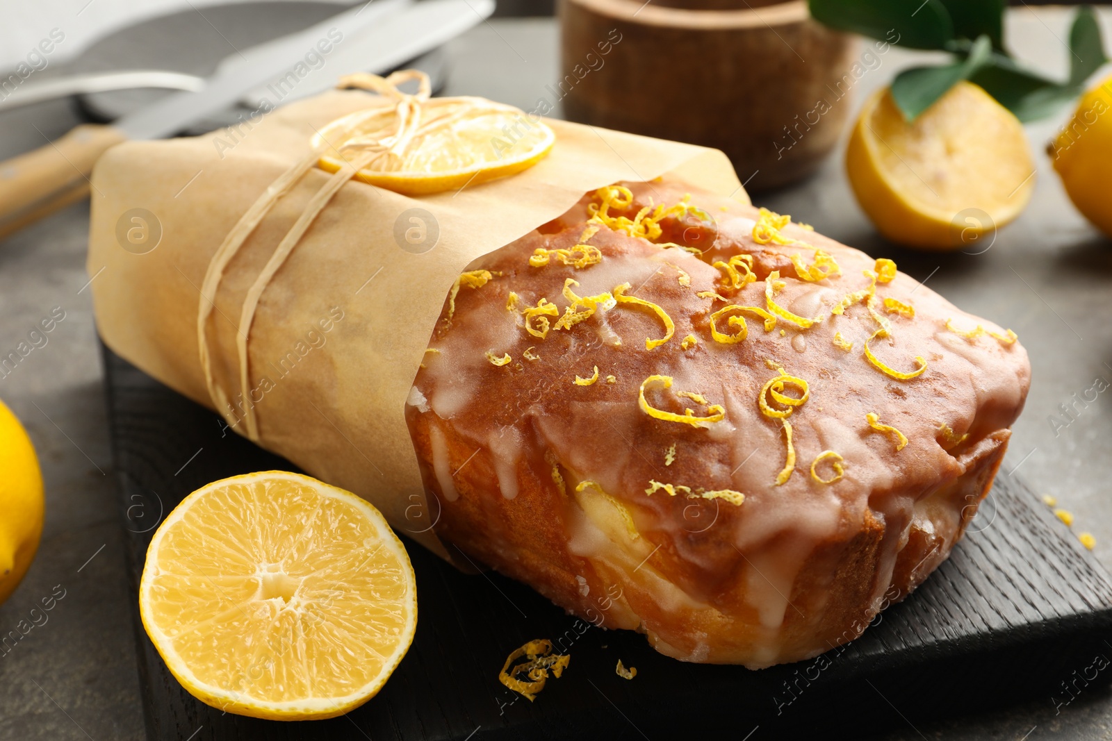 Photo of Wrapped tasty lemon cake with glaze and citrus fruits on grey table, closeup