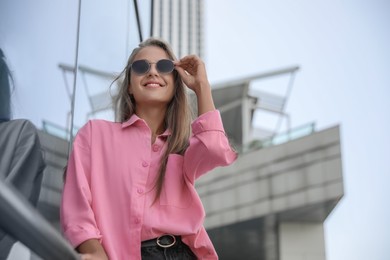 Beautiful young woman in stylish sunglasses near building outdoors, space for text