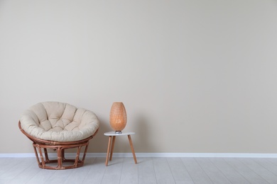Photo of Comfortable papasan chair and lamp on table near color wall with space for text. Interior element