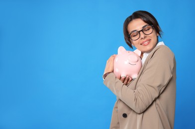 Young woman in eyeglasses with piggy bank on light blue background, space for text
