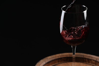 Photo of Pouring red wine into glass on wooden barrel against black background, closeup. Space for text