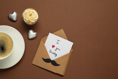 Photo of Father's day celebration. Card with phrase I Love You Daddy, cup of aromatic coffee, cupcake and heart shaped candies on brown background, flat lay. Space for text