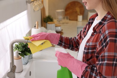 Photo of Woman with spray bottle and microfiber cloth cleaning water tap in kitchen, closeup