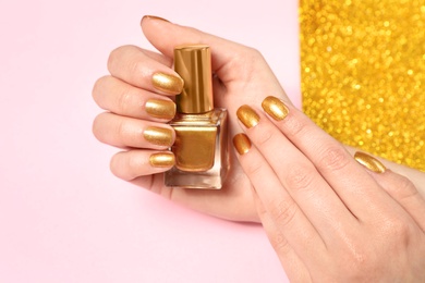 Photo of Woman holding bottle of golden nail polish in manicured hand on color background, closeup