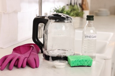 Photo of Cleaning electric kettle. Bottle of vinegar, sponge, rubber gloves and baking soda on countertop in kitchen