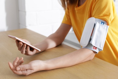 Photo of Woman checking blood pressure with modern monitor and smartphone at table indoors, closeup. Cardiology concept