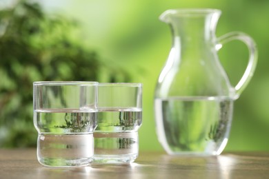 Photo of Jug and glasses with clear water on wooden table against blurred green background, closeup