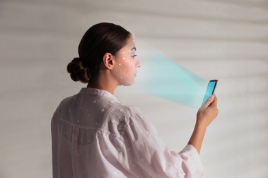 Image of Young woman unlocking smartphone with facial scanner near white wall. Biometric verification
