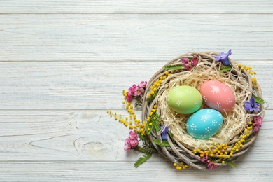 Photo of Wicker nest with painted Easter eggs and flowers on wooden table, top view. Space for text