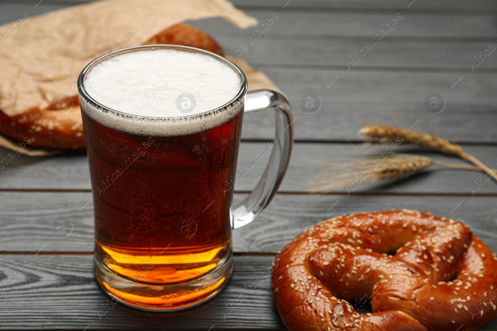 Photo of Mug of beer with tasty freshly baked pretzel on grey wooden table, closeup