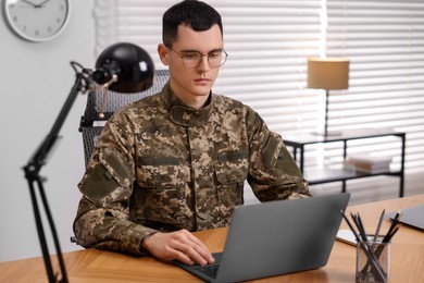 Photo of Military service. Young soldier working with laptop at wooden table in office