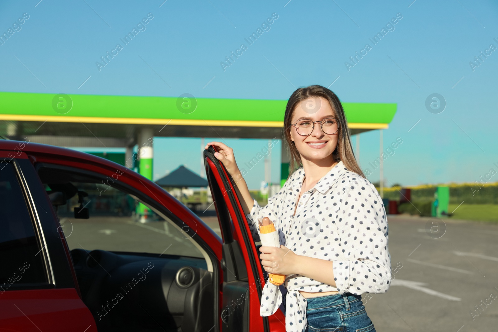 Photo of Beautiful young woman with hot dog opening car door at gas station