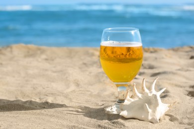 Glass of cold beer and seashell on sandy beach near sea. Space for text
