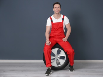 Young mechanic in uniform sitting on car tire near dark wall