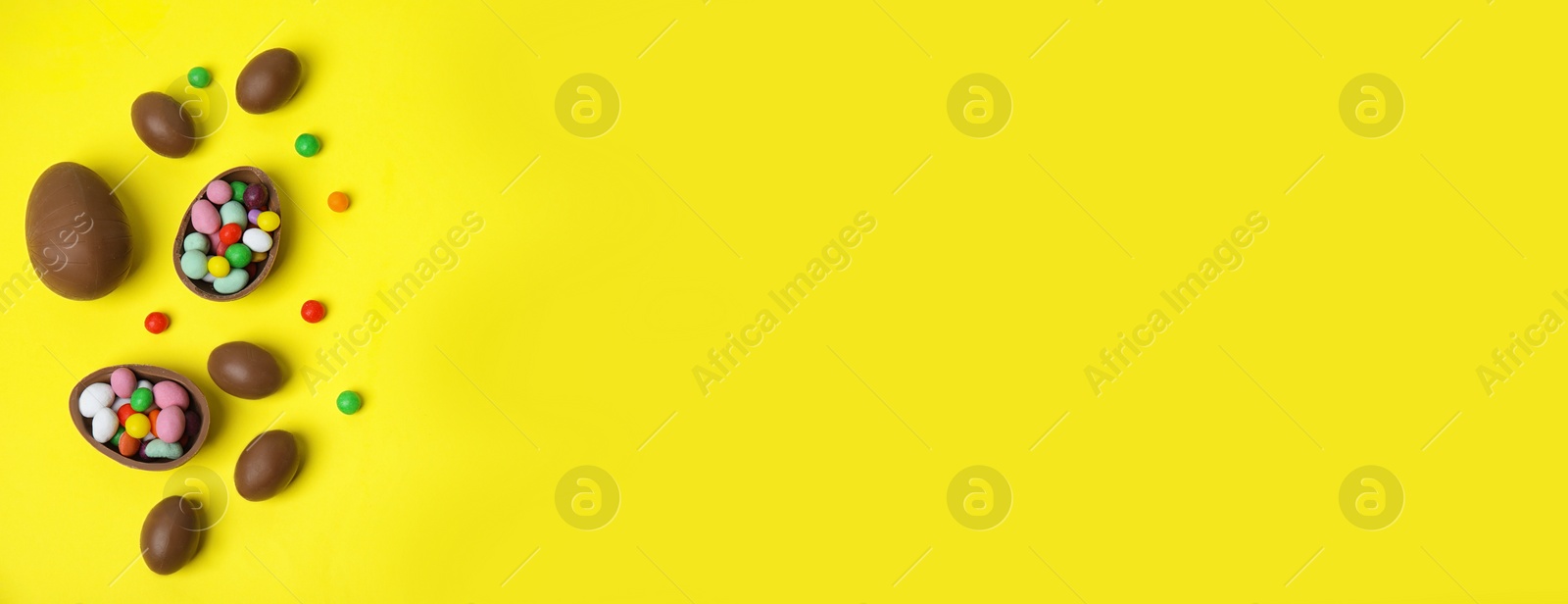 Image of Tasty chocolate eggs and colorful candies on yellow background, flat lay with space for text. Banner design