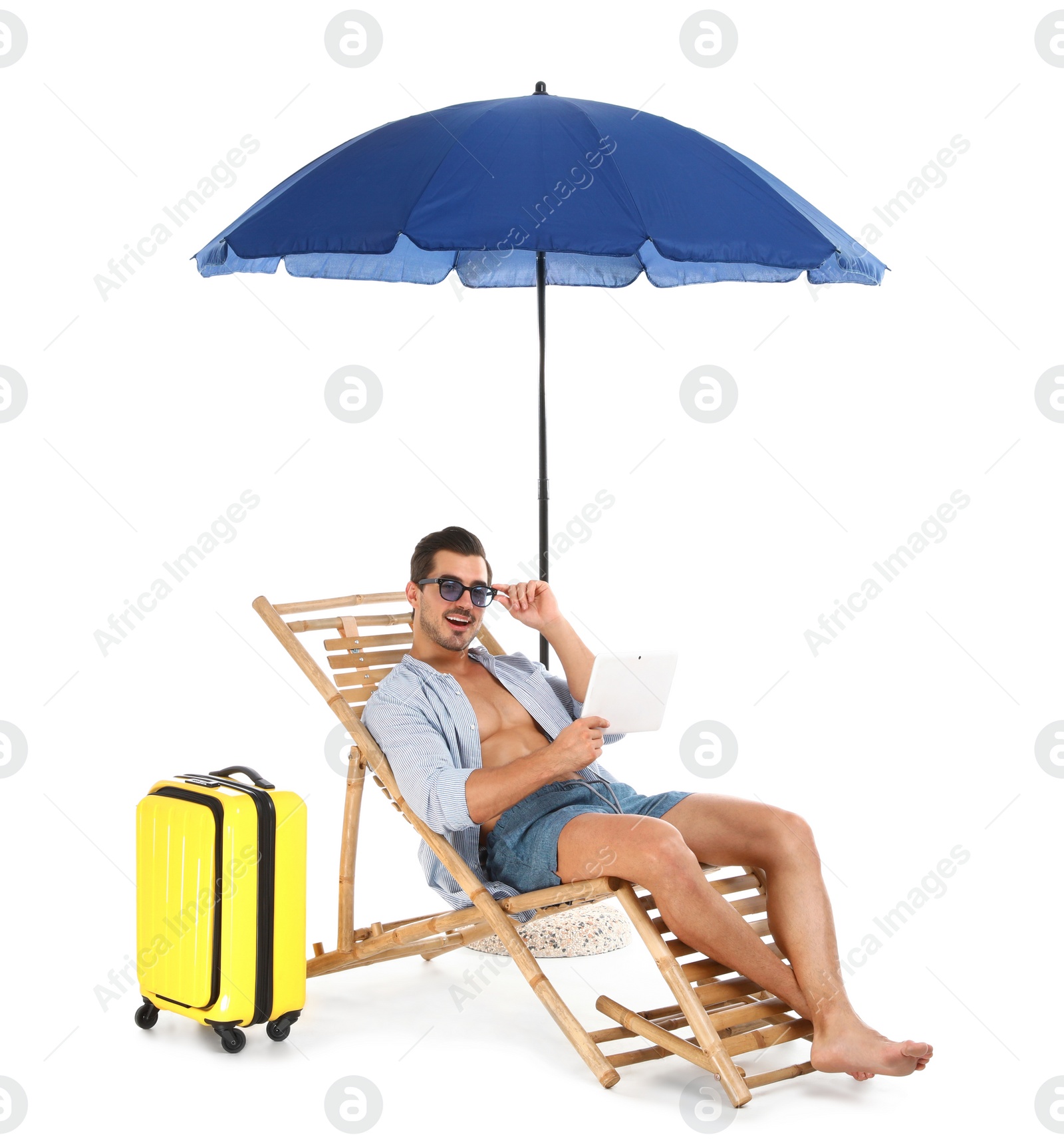 Photo of Young man with tablet and suitcase on sun lounger under umbrella against white background. Beach accessories