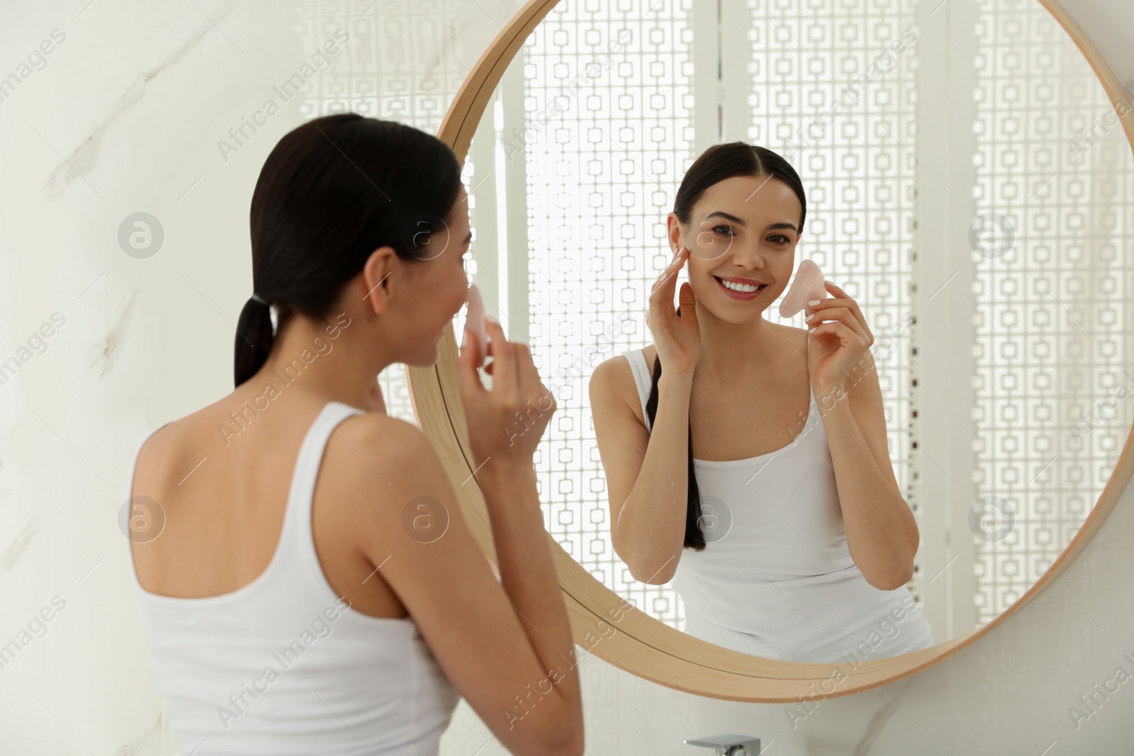 Photo of Beautiful young woman doing facial massage with gua sha tool in front of mirror at home