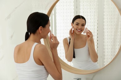 Photo of Beautiful young woman doing facial massage with gua sha tool in front of mirror at home