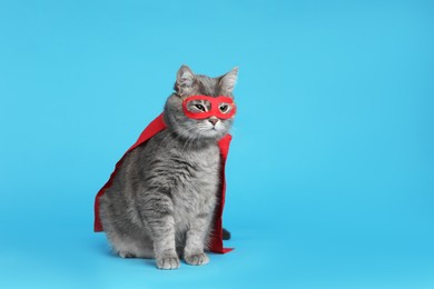 Photo of Adorable cat in red superhero cape and mask on light blue background, space for text