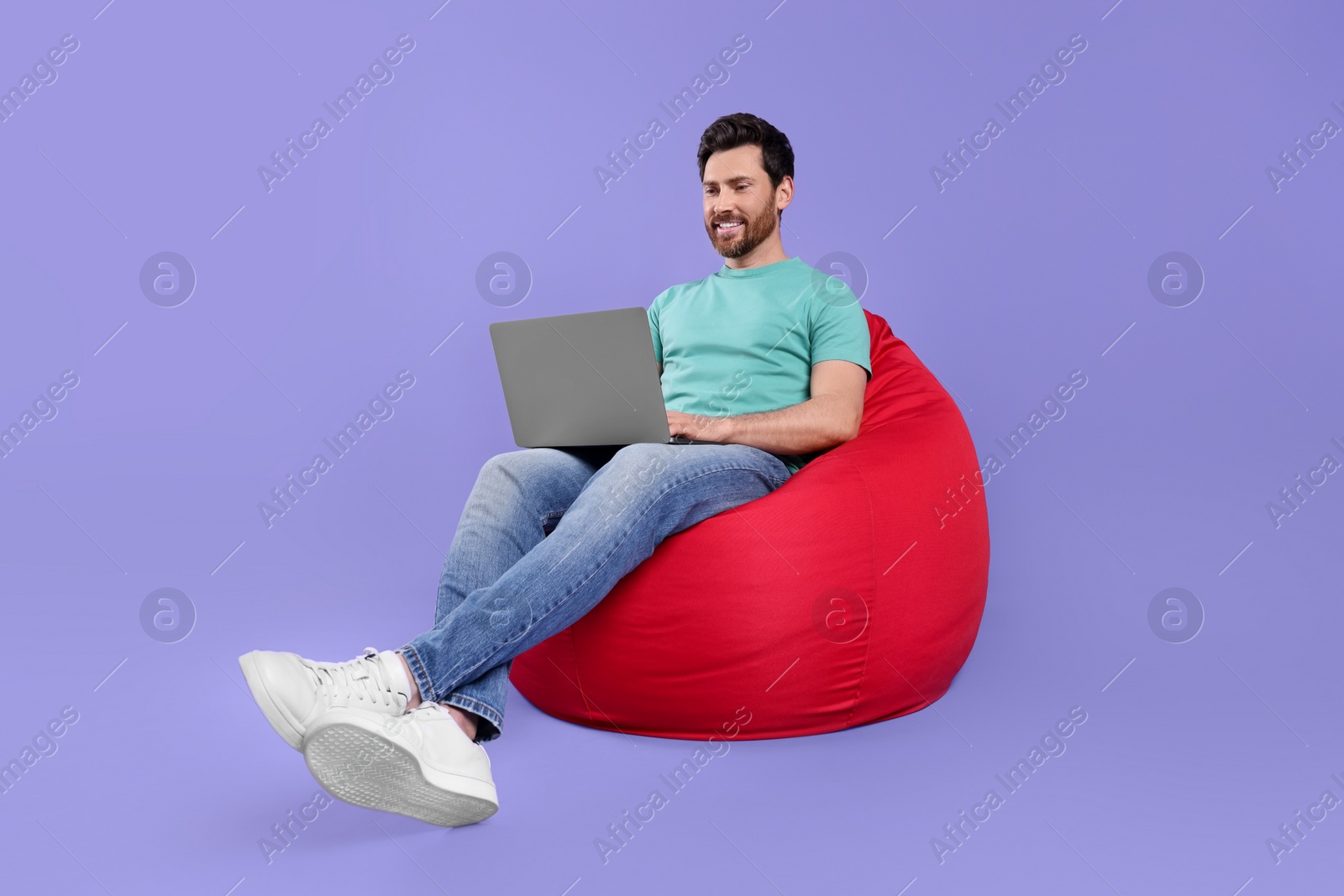Photo of Happy man with laptop sitting on beanbag chair against purple background