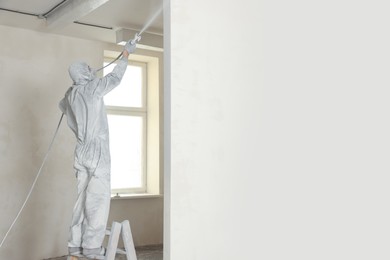 Decorator in uniform painting wall with sprayer indoors. Space for text