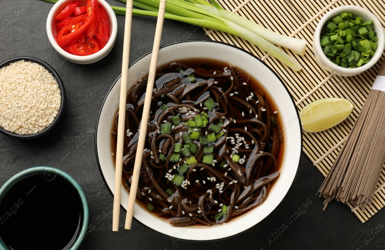 Photo of Tasty soup with buckwheat noodles (soba), ingredients and chopsticks on dark grey table, flat lay