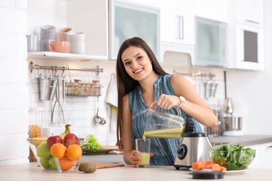 Young woman pouring tasty healthy smoothie into glass at table in kitchen