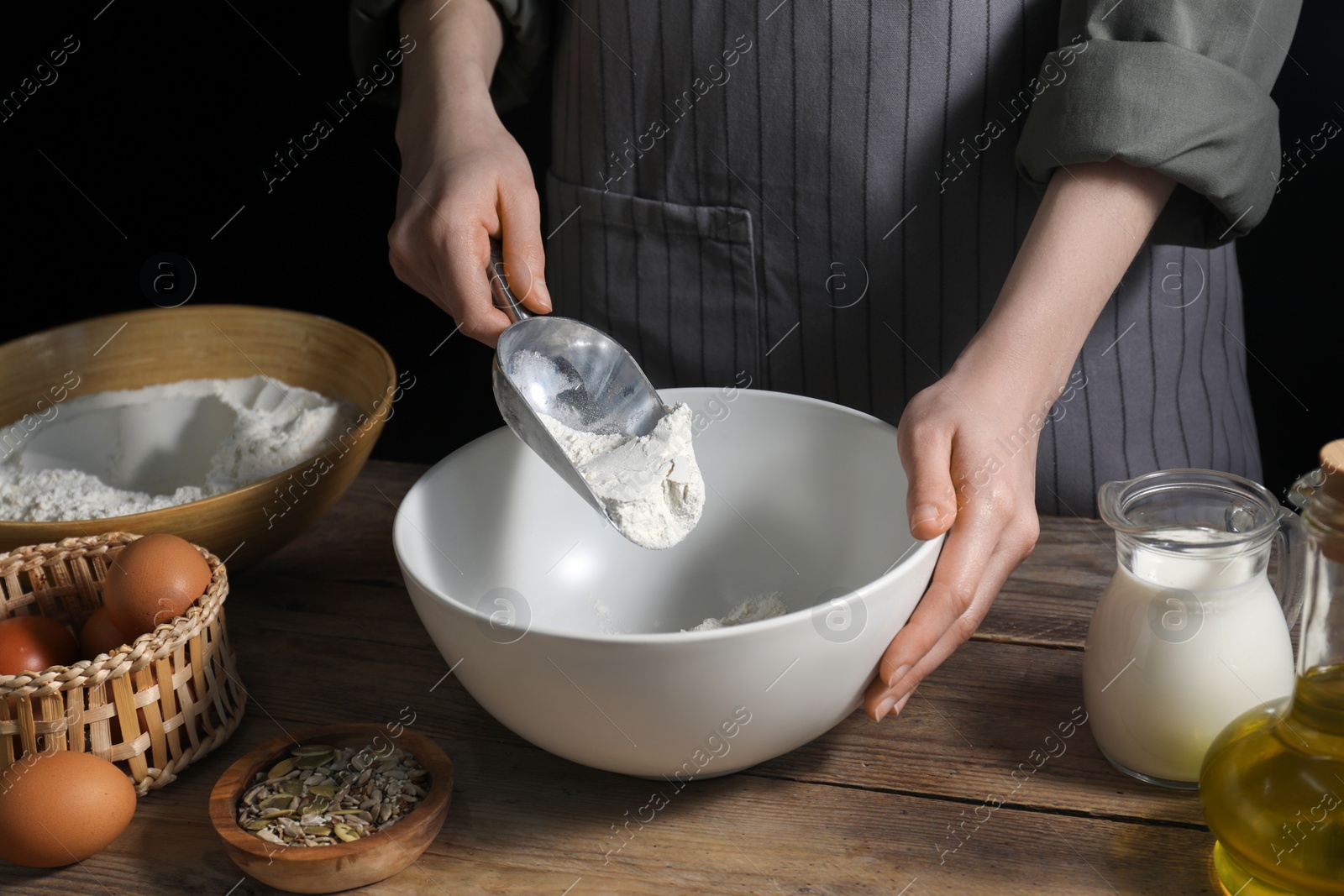 Photo of Making bread. Woman adding flour into bowl at wooden table on dark background, closeup