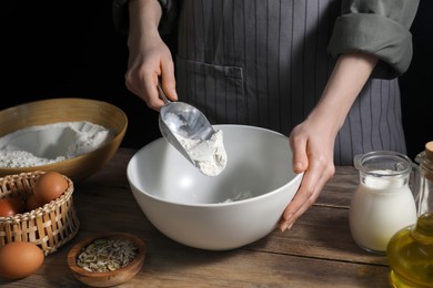 Making bread. Woman adding flour into bowl at wooden table on dark background, closeup