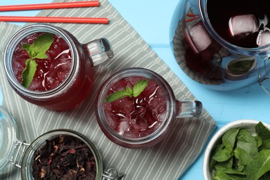 Photo of Delicious iced hibiscus tea with mint and dry flowers on light blue wooden table, flat lay