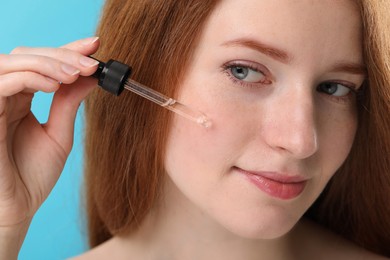 Beautiful woman with freckles applying cosmetic serum onto her face against light blue background, closeup