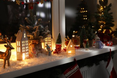 Photo of Christmas atmosphere. Beautiful glowing houses, fir trees, artificial snow and toys on window sill indoors