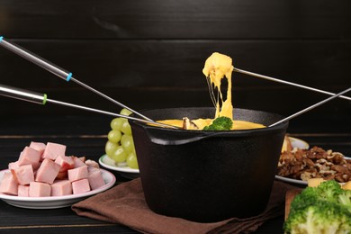 Dipping different products into fondue pot with melted cheese on black wooden table, closeup