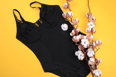 Photo of Elegant black plus size women's underwear and cotton flowers on yellow background, flat lay