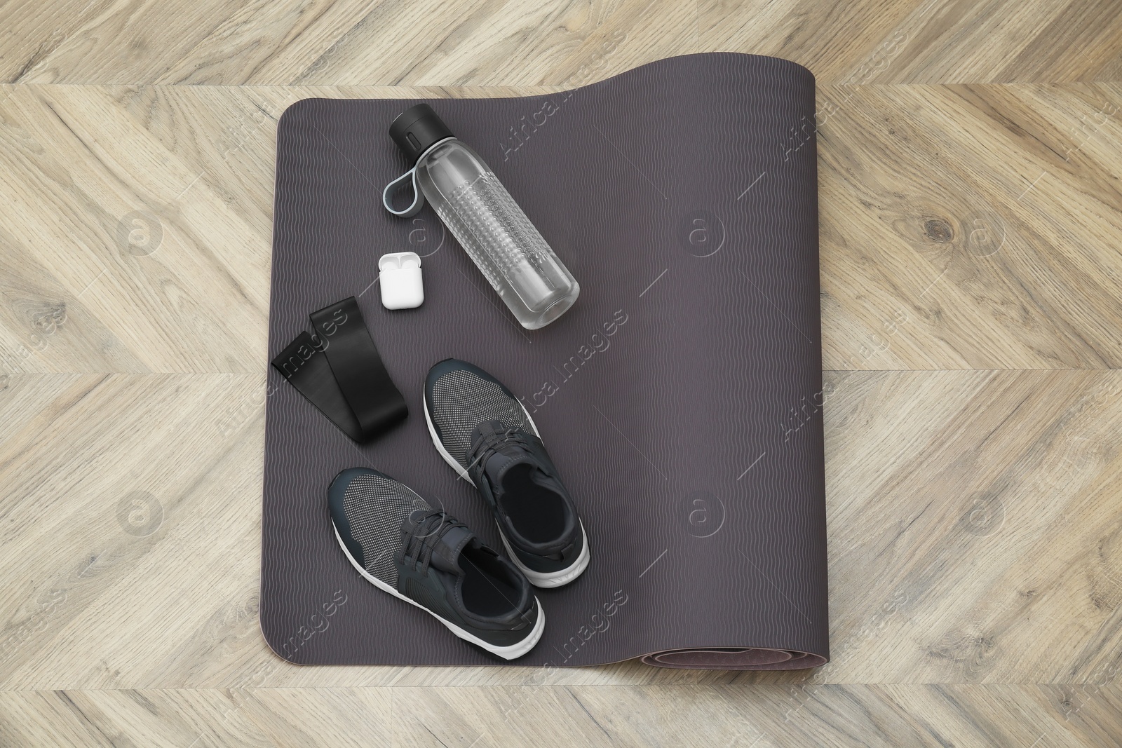 Photo of Exercise mat, bottle of water, wireless earphones, fitness elastic band and shoes on wooden floor, top view
